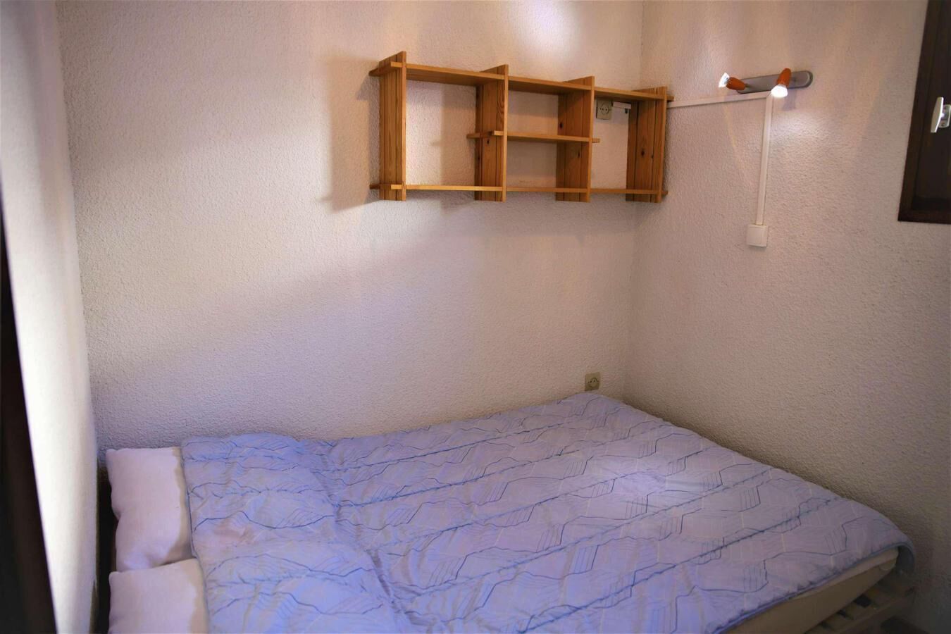 A small, single-bed room with a plain blue mattress and a wooden shelf unit above it.