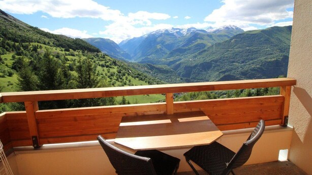 A balcony with a wooden railing, a table, and two chairs, overlooking a mountainous landscape with green valleys and snow-capped peaks. - View open, balcony, parking, tv, ski locker, 27m², Auris en Oisans (Auris)