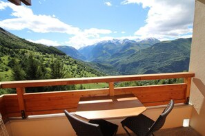 A balcony with a wooden railing, a table, and two chairs, overlooking a mountainous landscape with green valleys and snow-capped peaks. - View open, balcony, parking, tv, ski locker, 27m², Auris en Oisans (Auris)