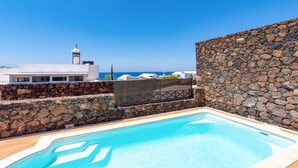 Une piscine bordée d’un mur de pierre, un bâtiment blanc et une vue sur la mer.
