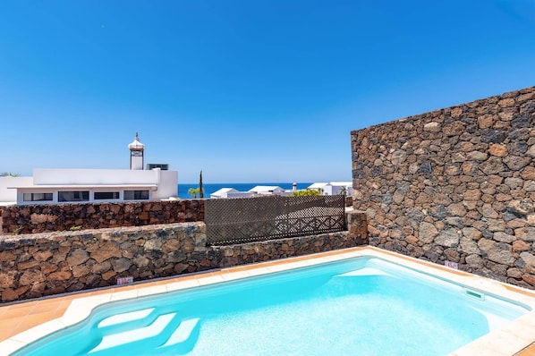 Une piscine bordée d’un mur de pierre, un bâtiment blanc et une vue sur la mer.