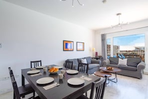 Un salon moderne avec un coin repas, des canapés gris et une grande fenêtre donnant sur un beau paysage.