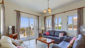 A spacious living room with a sofa, armchair, and a table. Large windows offer a view of the outdoors. - Villa Camelia by Villa Plus (Peyia)