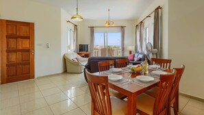 A dining area with a table set for four, a sofa, and a TV in the background. - Villa Camelia by Villa Plus (Peyia)