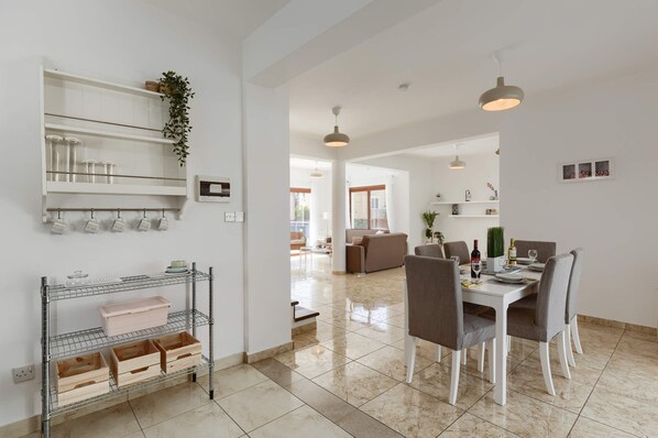 A modern kitchen with a dining area, featuring a metal rack with storage containers, a dining table set for four, and a view into the living room.