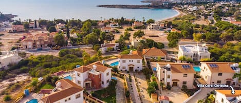 A coastal residential area with villas, including Villa Coral Christina, featuring terraced roofs, swimming pools, and a view of the sea.