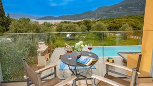 Un balcon avec une table dressée pour deux personnes, donnant sur une piscine et offrant une vue panoramique sur les montagnes et un plan d’eau.