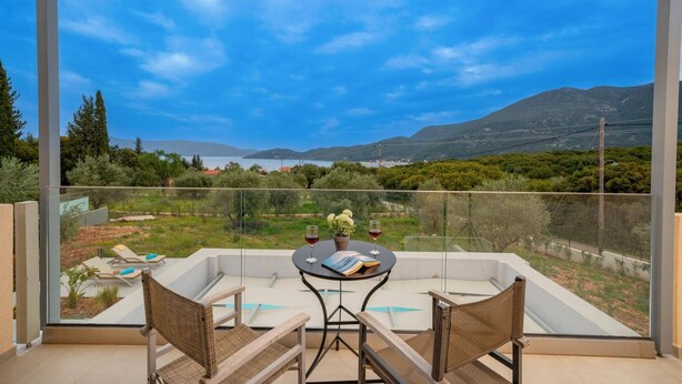 Un balcon doté d’une paroi vitrée, deux fauteuils en osier, une petite table ronde avec un livre et deux verres à vin, et une vue sur un paysage montagneux avec un plan d’eau.