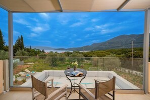 Ein Balkon mit Glasveranda, zwei Korbstühle, ein kleiner runder Tisch mit einem Buch und zwei Weingläsern sowie Blick auf eine Berglandschaft mit einem Gewässer.