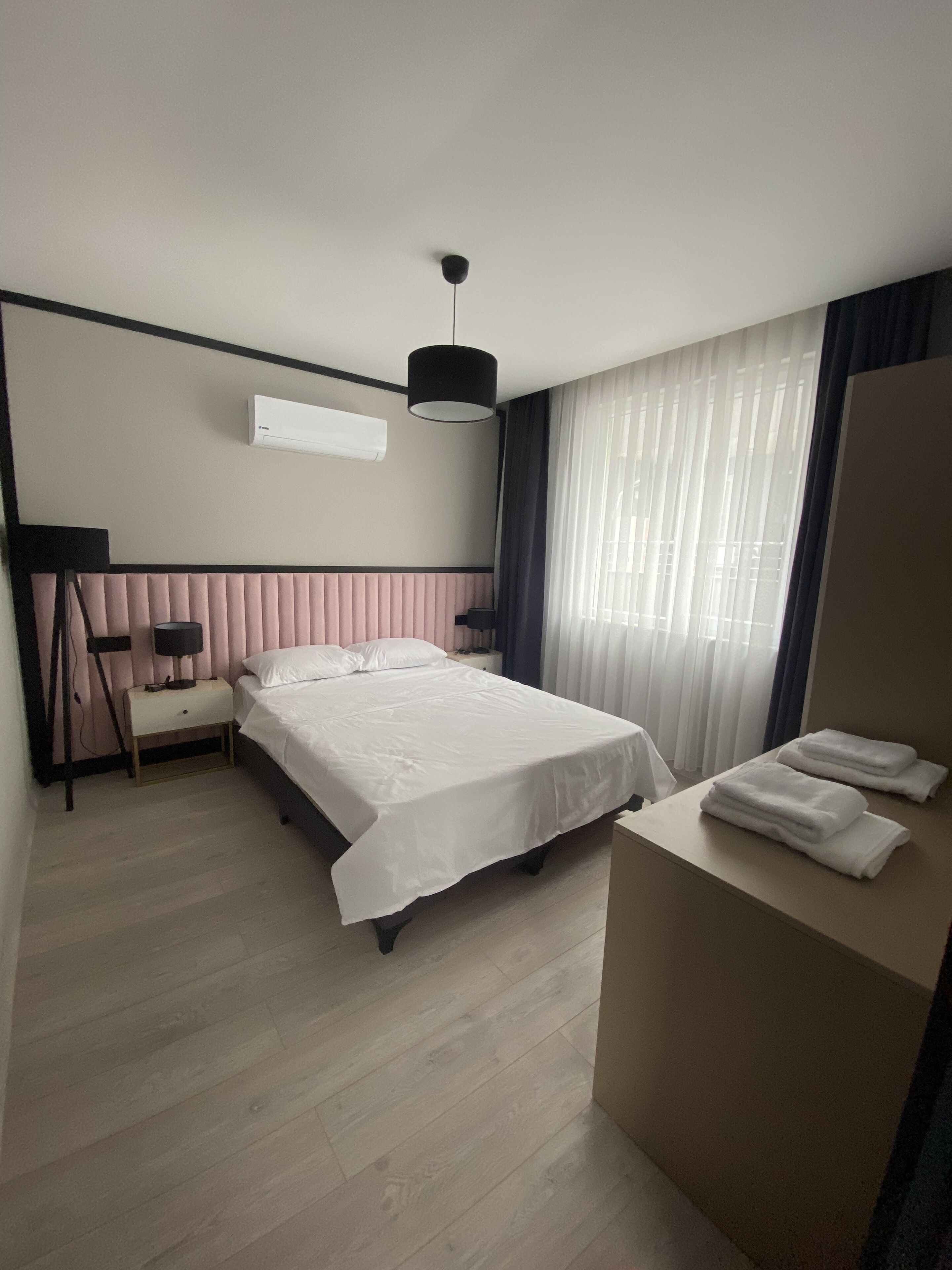 A hotel room with a bed, bedside table, lamp, air conditioning unit, and a window with curtains.