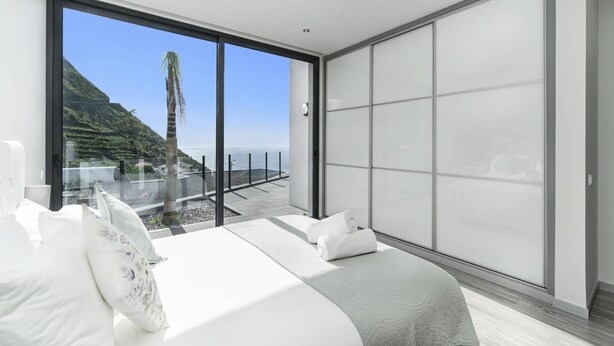 A modern bedroom with a large sliding glass door leading to a balcony with a scenic view of a coastal landscape. - Villa Calheta Charm by Villa Plus (Arco Da Calheta)