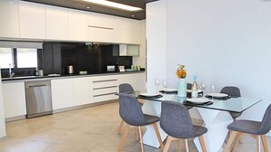A modern kitchen with white cabinets, a dishwasher, and a dining area with a glass table and four grey chairs. - Villa Calheta Charm by Villa Plus (Arco Da Calheta)