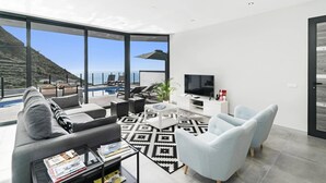 A modern living room with a large window overlooking a pool and a scenic view. - Villa Calheta Charm by Villa Plus (Arco Da Calheta)