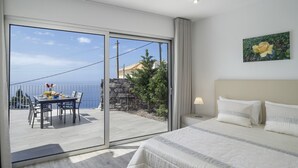 Habitación de hotel con cama, mesita de noche, mesa con sillas y vista al mar a través de una puerta corrediza de vidrio.