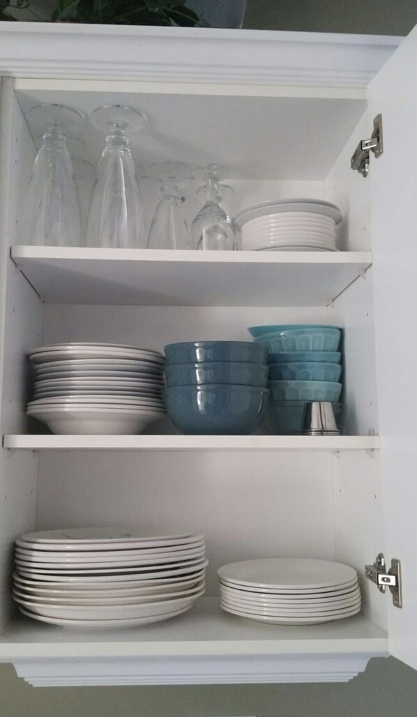 A white cabinet with multiple shelves containing stacks of white plates, blue bowls, and clear glass bottles.