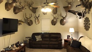 A living room with a brown leather sofa, a flat-screen TV mounted on the wall, and a ceiling fan. - Outdoorsman Paradise- 1/2 mile from Lake Oahe (Pierre)