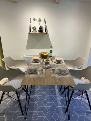 A dining area with a wooden table set for four, surrounded by modern chairs. A shelf above holds decorative items and a vase.