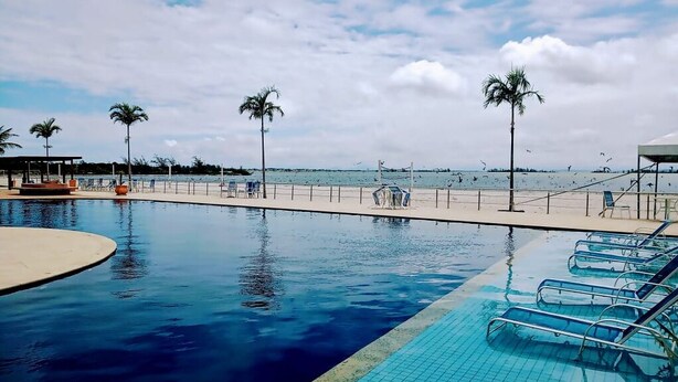 A swimming pool with lounge chairs, palm trees, and a beach in the background. - Golden Lake Residence (Arraial do Cabo)