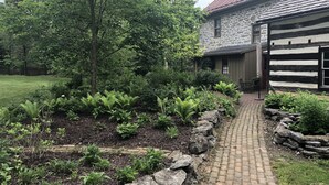 Una casa de pedra amb un porxo de fusta, envoltada per un jardí ple de frens i plantes florides, i un camí de maons que condueix a l'entrada.