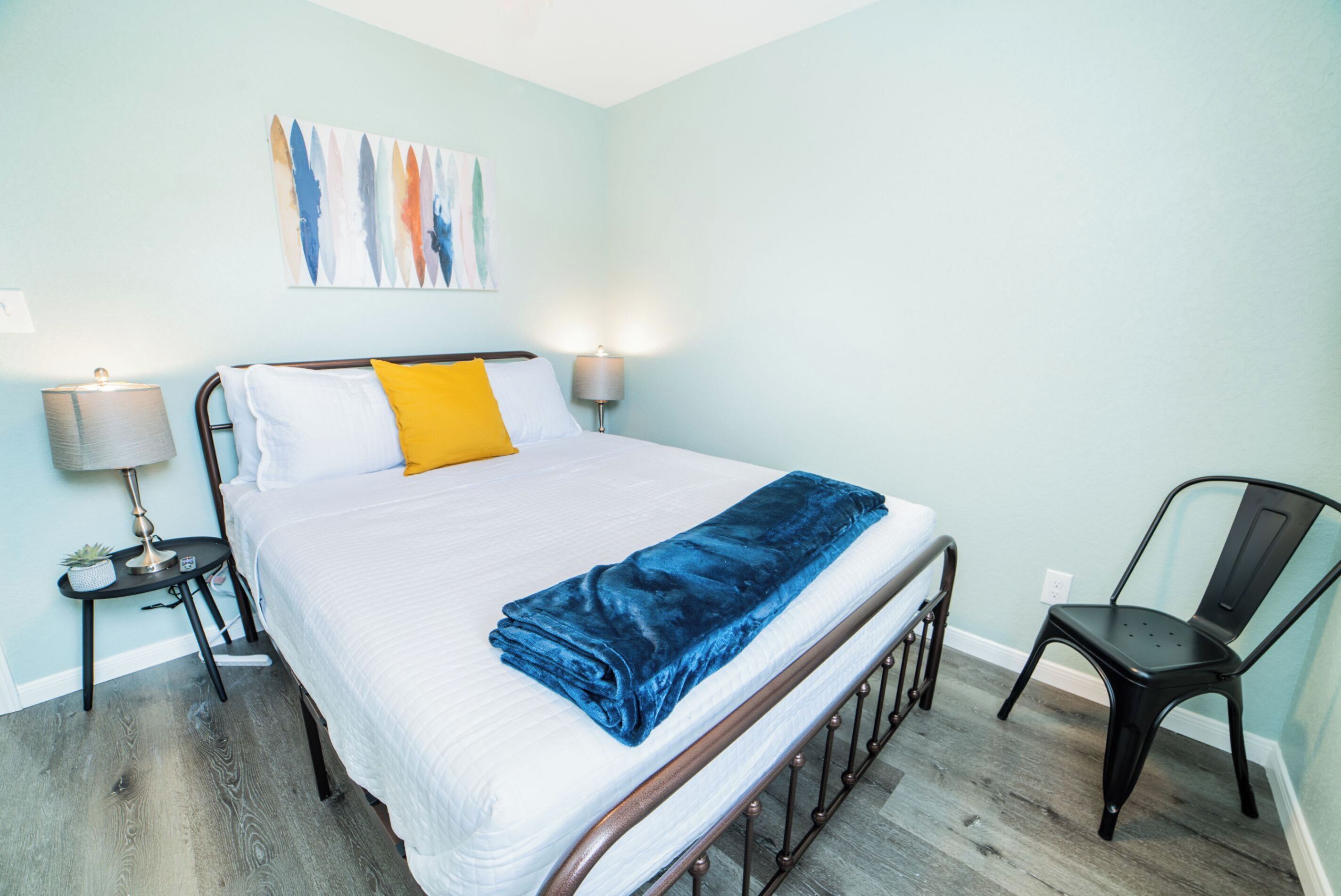 A neatly made bed with white bedding and a blue blanket, two bedside lamps, a small table with a lamp, a chair, and a wall-mounted abstract painting.
