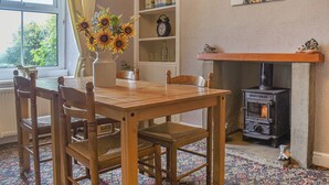 A cozy dining area with a wooden table and chairs, a vase of sunflowers, a bookshelf, a clock, and a fireplace with a wood-burning stove. - Bondcroft Farm Cottage (Eastby, near Skipton)