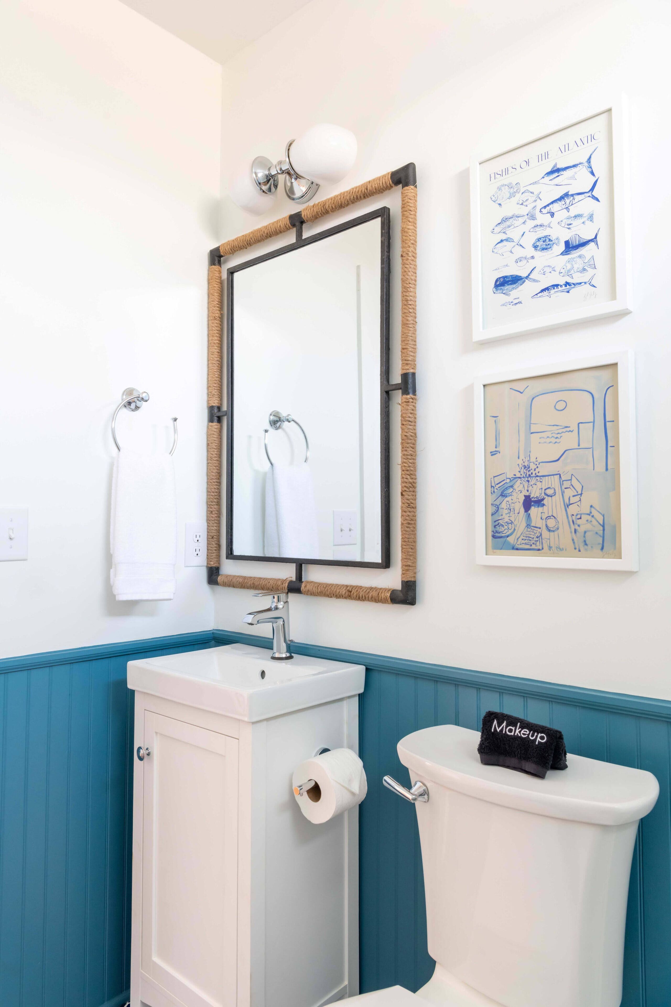 A bathroom with a white sink, a toilet, and a mirror with a wooden frame. There are two towels hanging on hooks and two framed artworks on the wall.