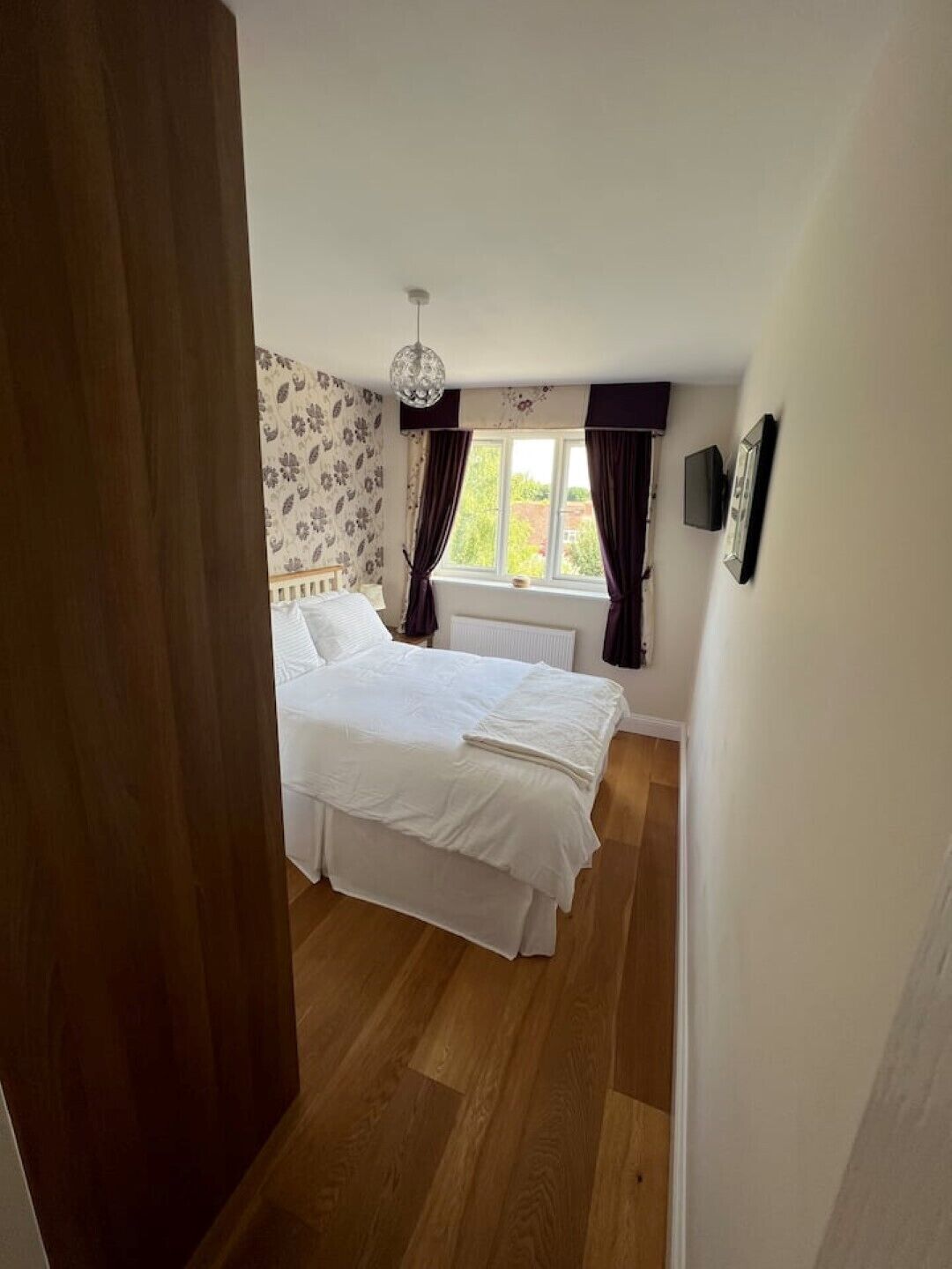 Une chambre à coucher avec un lit, une fenêtre avec des rideaux, un radiateur et un téléviseur fixé au mur.