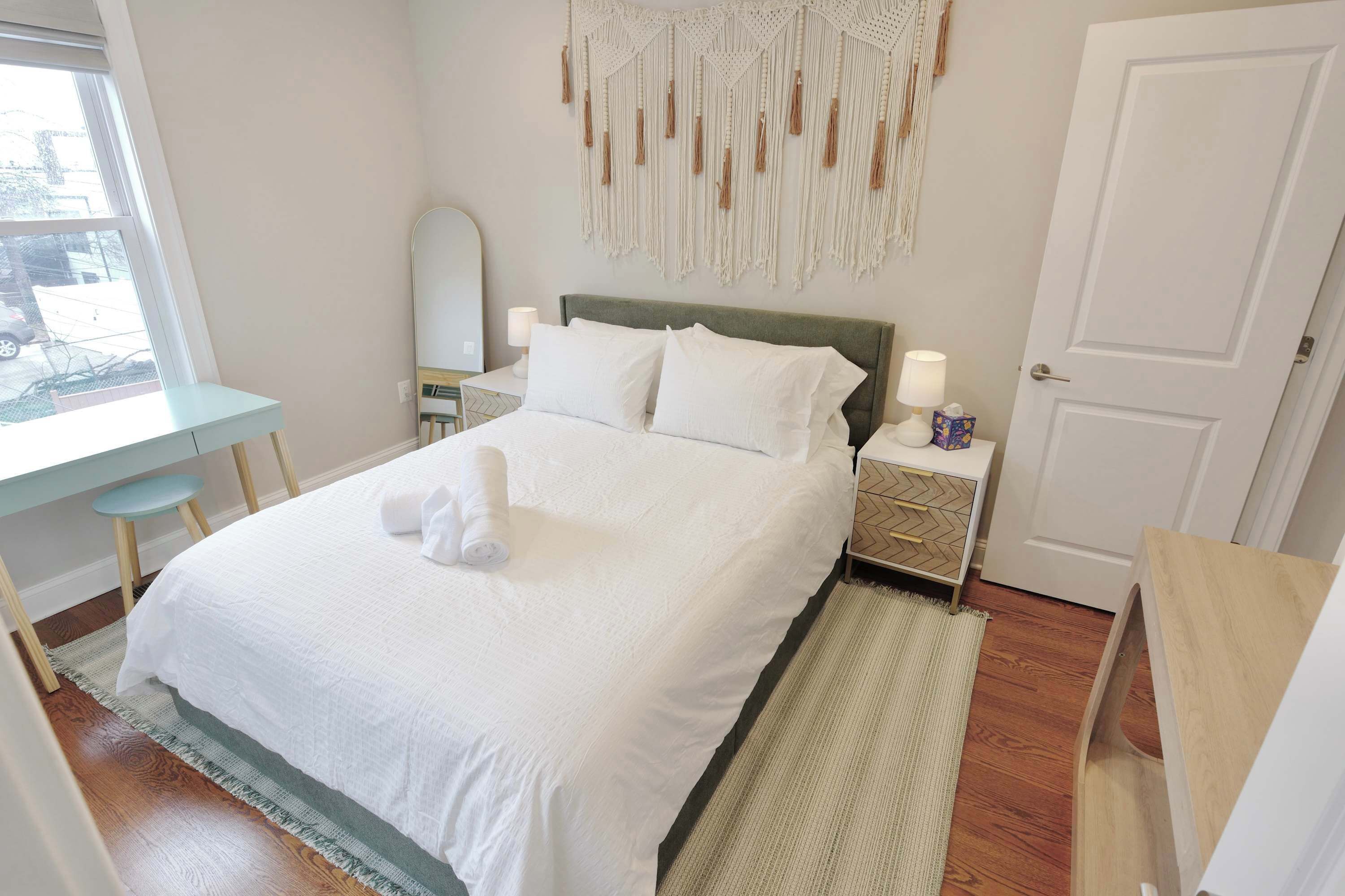 Un letto rifatto con lenzuola bianche, un comodino con una lampada, un pannello decorativo in macramè a parete e una finestra con vista all'esterno.