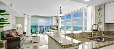 A modern kitchen with a large island, a dining area with a view, and a living room with a sofa and a plant.