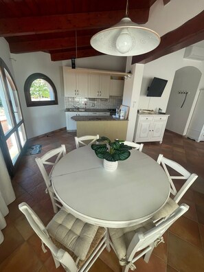 Un coin repas avec une table ronde, des chaises blanches, une plante en pot, une cuisine en arrière-plan et un téléviseur fixé au mur.
