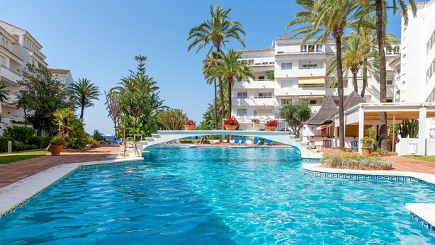 En swimmingpool med hvid poolterrasse, palmer og en bygning med balkoner i baggrunden.
