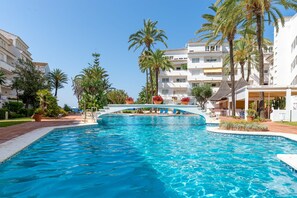 Une piscine avec une terrasse blanche, des palmiers et un bâtiment doté de balcons en arrière-plan.