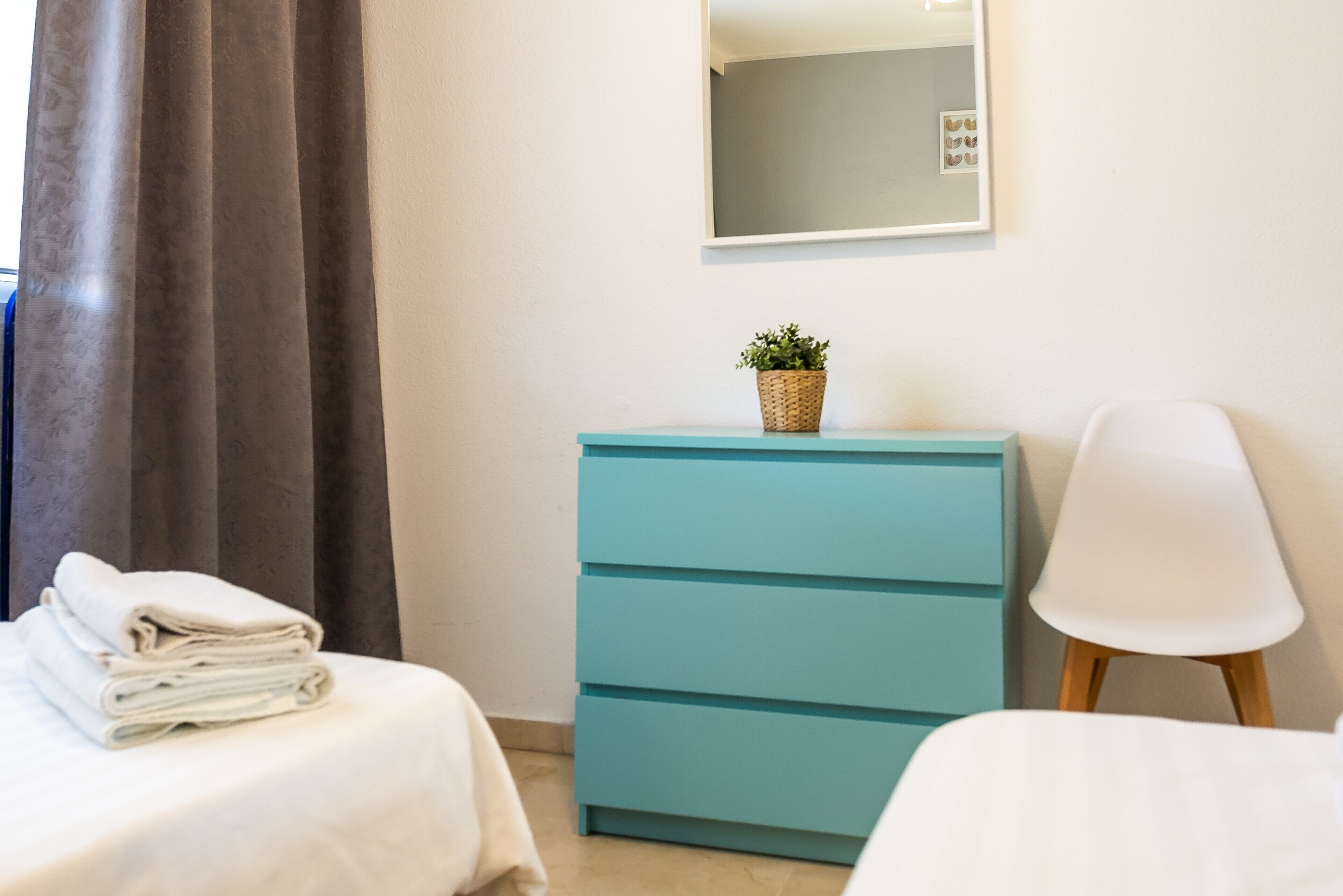 A hotel room with a bed, a blue dresser, a white chair, and a small potted plant on the dresser.