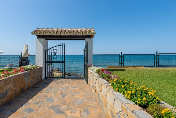 Un chemin de pierres menant à une pergola agrémentée d’une porte, entourée de parterres de fleurs et offrant une vue sur la mer.