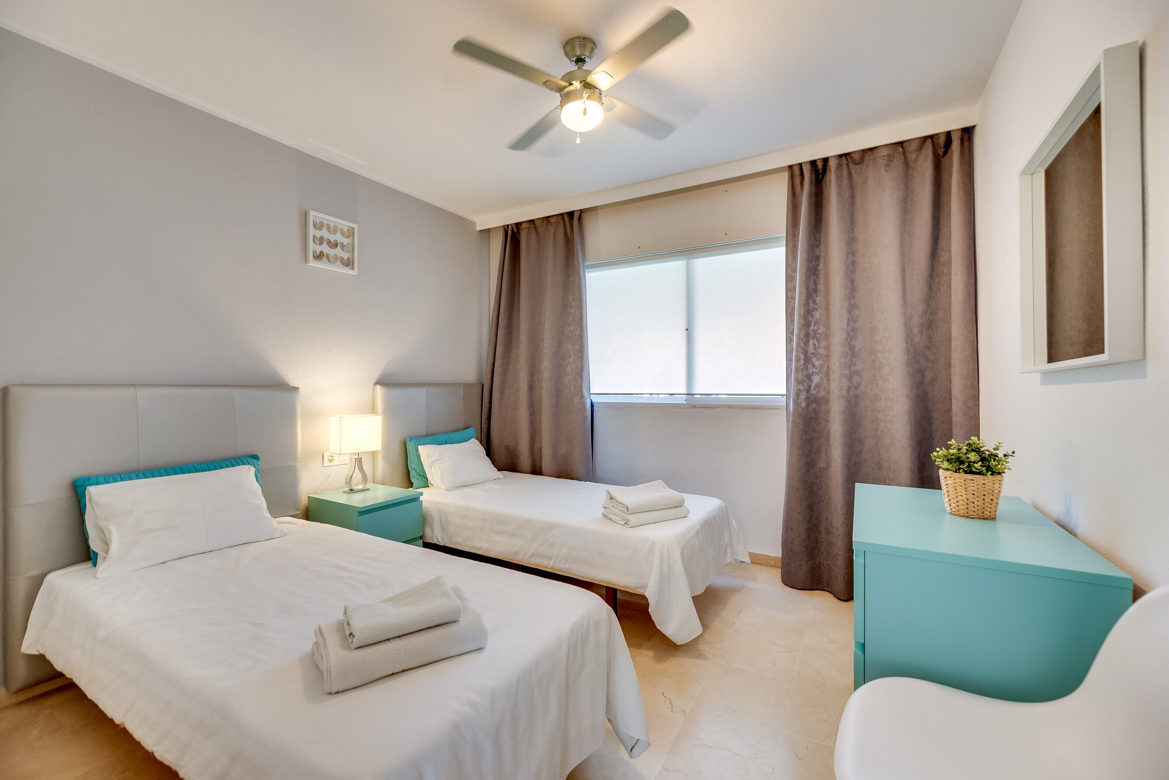 Una habitación de hotel con dos camas, un ventilador de techo, una ventana con cortinas, una mesita de noche con una lámpara y una planta pequeña en una mesita auxiliar.