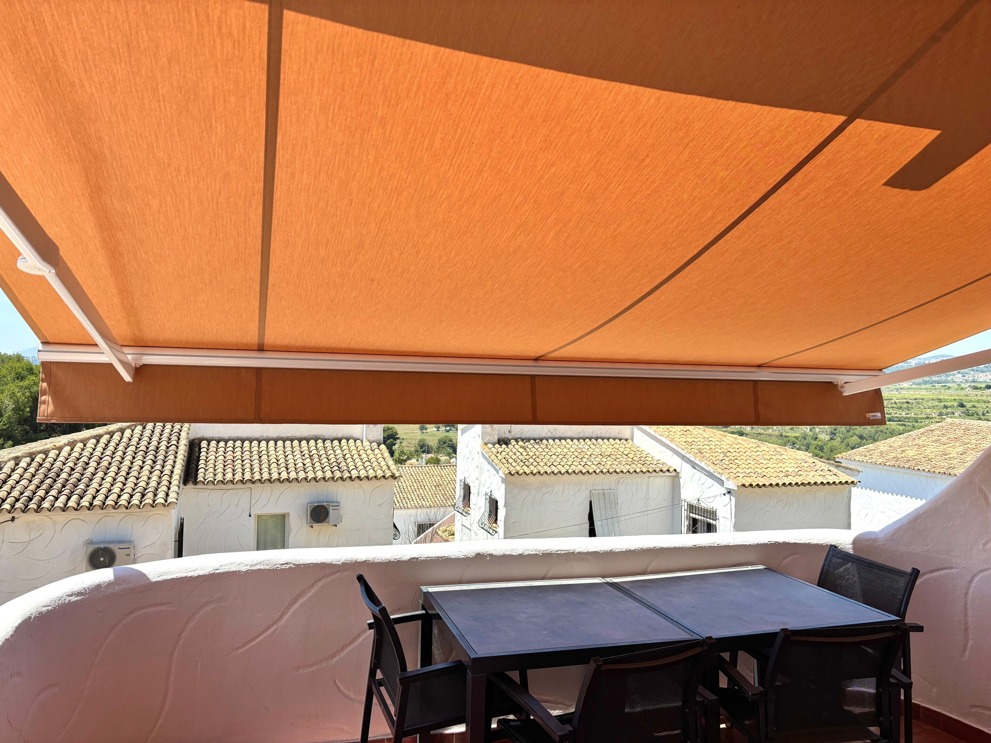 Een balkon met een tafel en stoelen, uitzicht op gebouwen met terracotta daken, en een grote oranje luifel.