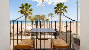 Een balkon met een tafel en stoelen, met uitzicht op een strand met palmbomen en mensen.