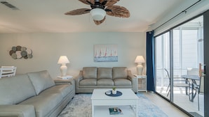 A living room with a sofa, coffee table, and two lamps. There is a ceiling fan, a painting of sailboats, and a view through a glass door.