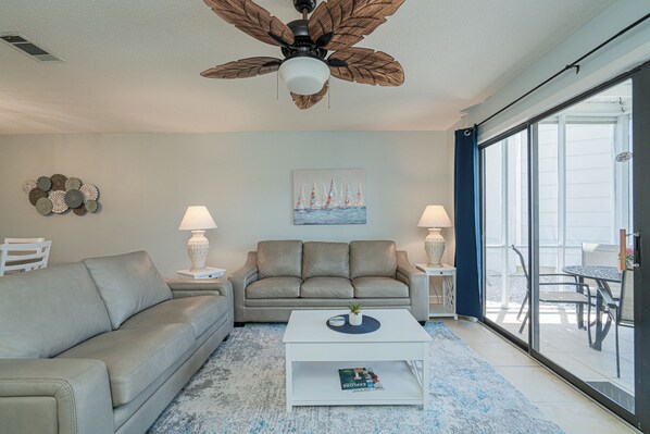 A living room with a sofa, coffee table, and two lamps. There is a ceiling fan, a painting of sailboats, and a view through a glass door. - Miramar Beach Townhouse - 3 Block Walk to Amazing Beach, 2 BR 2.5 BA, Sleeps 8 (Miramar Beach)