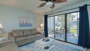 A living room with a sofa, coffee table, and a ceiling fan. There is a painting on the wall and a view of the outdoors through a glass door.