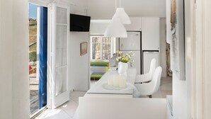 A modern kitchen with white cabinetry, a dining table with white chairs, a TV mounted above the counter, and a view of the outdoors through a door.