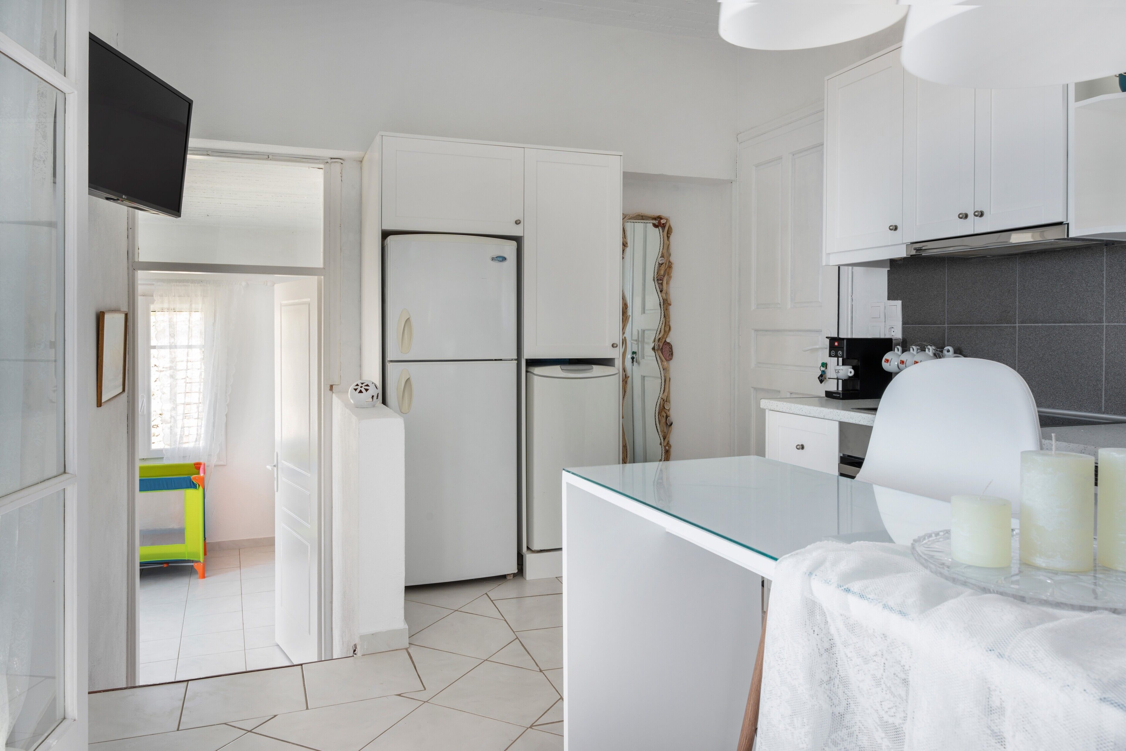 A kitchen with white cabinets, a refrigerator, a glass table with candles, and a view into a room with a bed and a window.