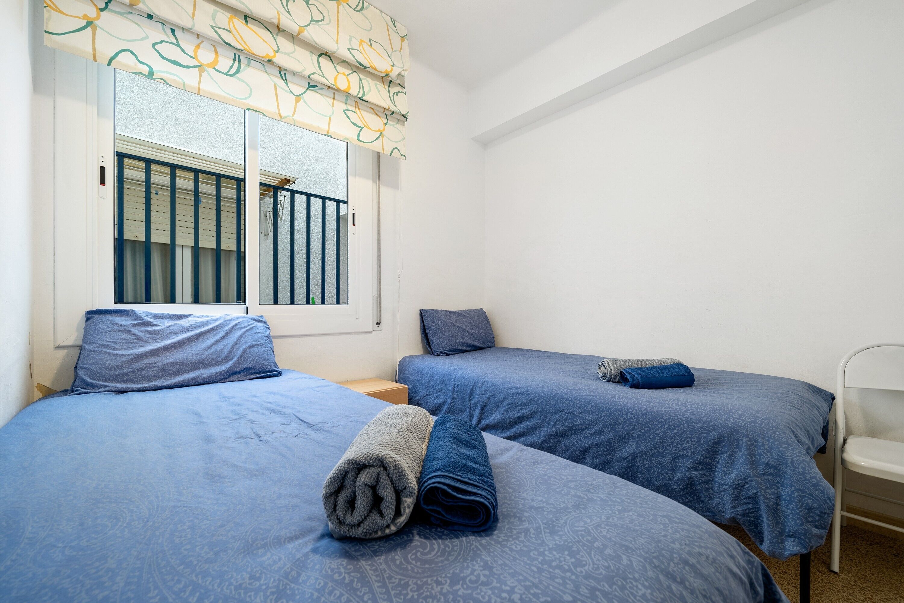 Una habitación con dos camas, una ventana con cortina, una silla y una mesita.