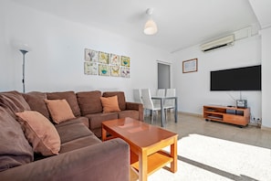 A living room with a brown sofa, a wooden coffee table, a TV, and a dining area with chairs and a table. - Apartment 'Vista Mar' with Sea View, Wi-Fi and Air Conditioning (l'ampolla)