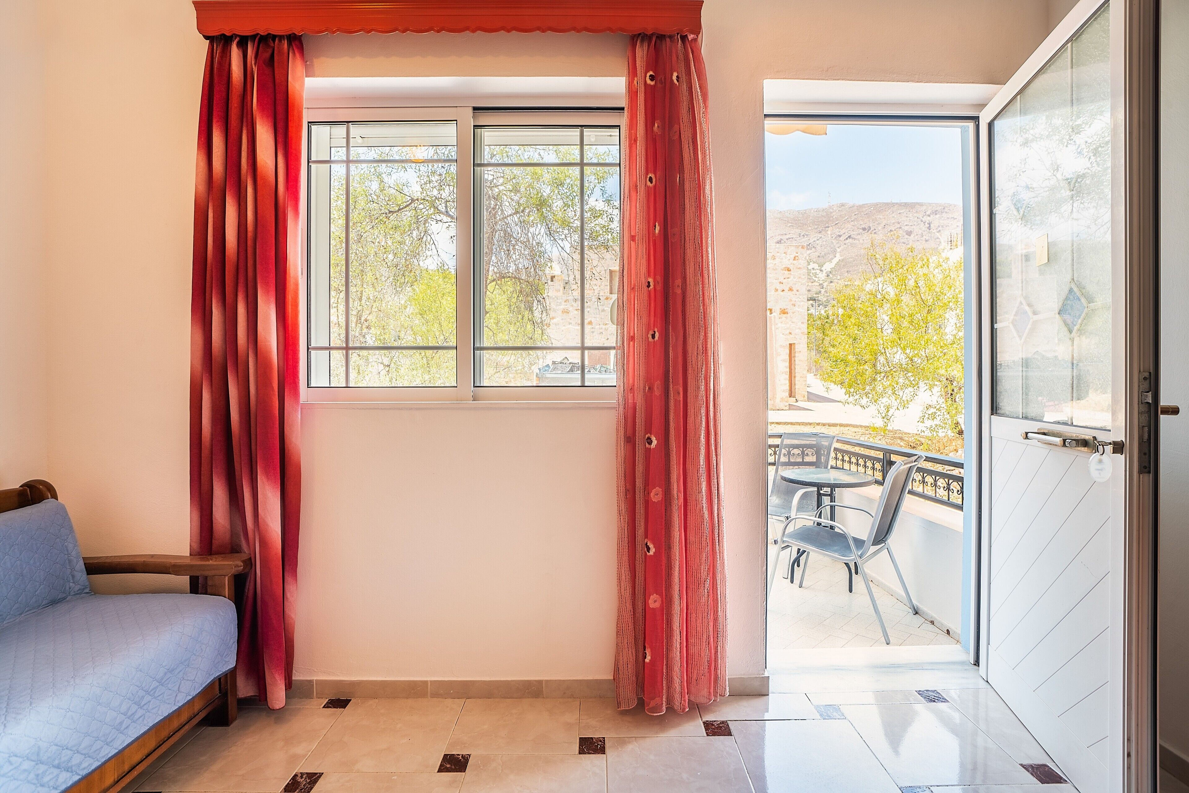 A room with a bed, a window with curtains, a door leading to an outdoor area with a table and chairs.