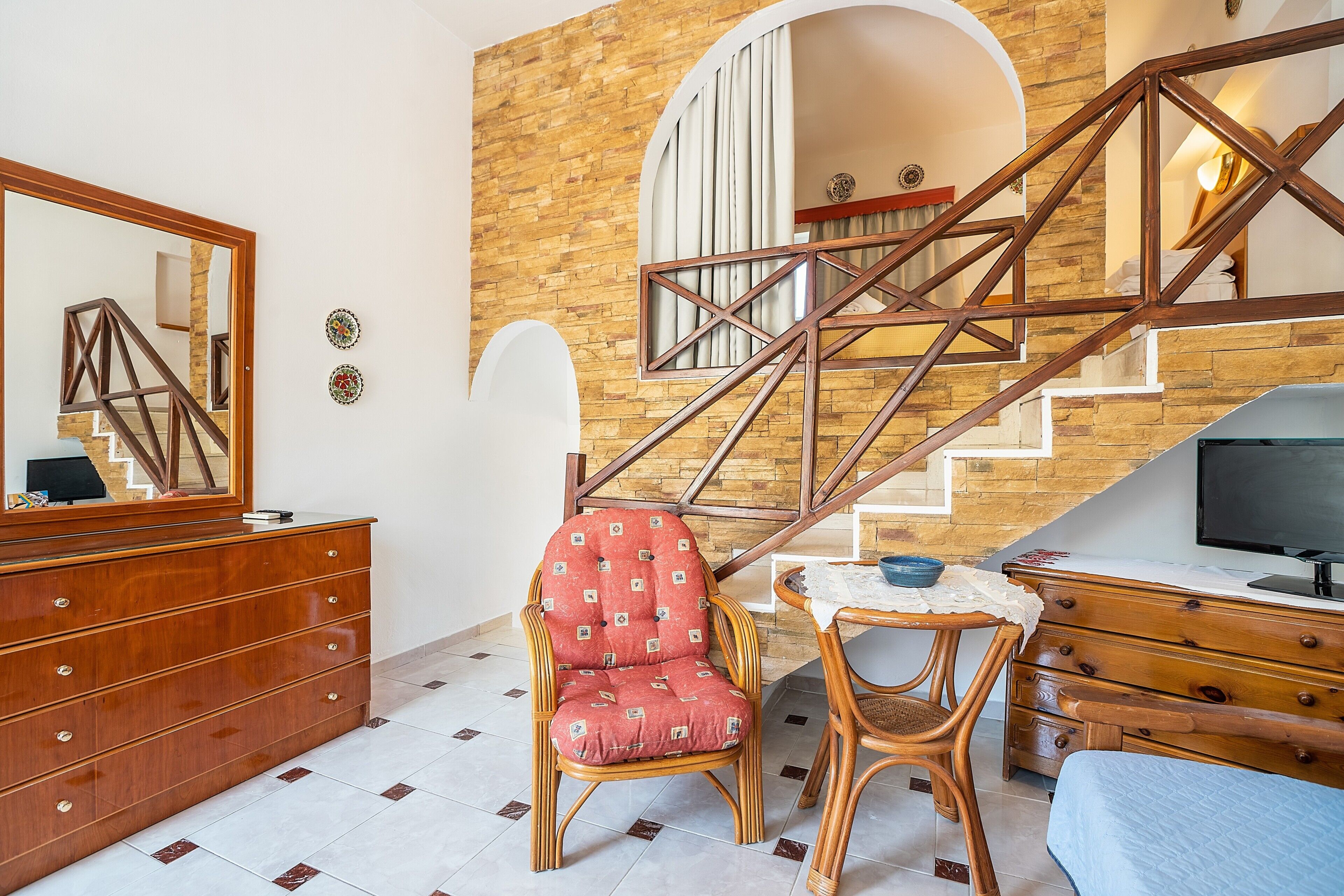 Une pièce avec un escalier en bois, une commode en bois, un fauteuil rouge avec coussin, une petite table recouverte d’une nappe en dentelle et un téléviseur.