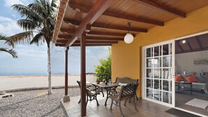 A covered patio with a table and chairs, a palm tree, and a view of the beach.