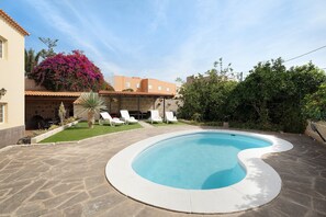 A swimming pool with a curved edge, surrounded by a stone patio. There are lounge chairs and a small lawn area with a palm tree.