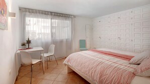 A bedroom with a bed, a desk, a chair, a window with sheer curtains, and a wardrobe with multiple doors.