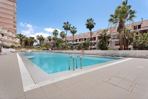 A swimming pool with two metal ladders, surrounded by palm trees and a building with balconies.
